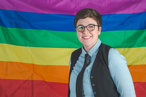 There is a young adult female in her 20's standing in front of a pride flag in a studio