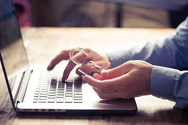 Closeup of man tying on a laptop keyboard while holding on one hand a lock like transparent object. Focus is on the lock symbol meaning online information security