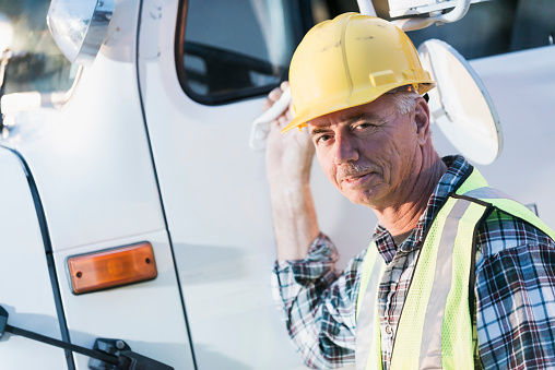 A mature man wearing a hardhat and safety vest standing outside a white truck, reaching for the door handle as he looks at the camera.