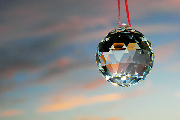 Faceted white quartz Feng Shui crystal ball hanging on red string with blue sky and pink and grey clouds on the background.