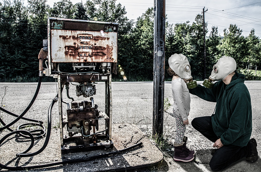 Father with gas mask placing mask on daughter besides destroyed gas pump during day