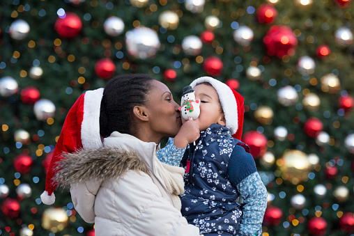 Mother and her child wearing red Santa hats, eating snowman candy, standing against Christmas tree outdoors.