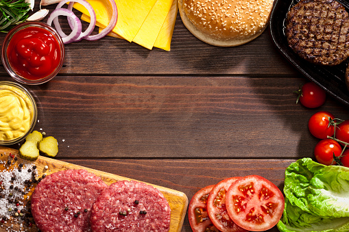 Ingredients for preparing delicious homemade cheeseburger placed all around a rustic wood table leaving a useful copy space at the center of the frame. Some of the ingredients included are: Ground meat, salt, pepper, pickles, tomato, onion, cheddar cheese, bacon, lettuce, sesame bread, ketchup and mustard.  DSRL studio photo taken with Canon EOS 5D Mk II and Canon EF 100mm f/2.8L Macro IS USM