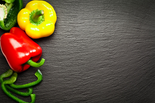 Top view of red, yellow and green bell peppers making a border at the left side of dark slate background leaving a useful copy space at the center-right of the frame. The green pepper is cut in half. DSRL studio photo taken with Canon EOS 5D Mk II and Canon EF 100mm f/2.8L Macro IS USM