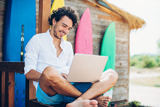 Surfing Cheerful young man using laptop with surfing boards at the background. pretty beach house stock pictures, royalty-free photos & images