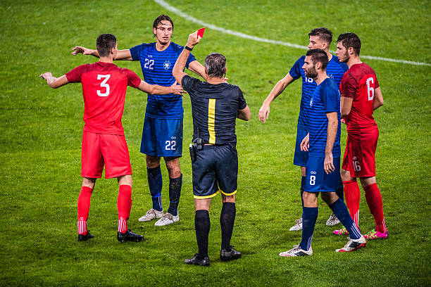 Referee holds up red card Referee holds up red card during football match, players arguing with referee. referee stock pictures, royalty-free photos & images