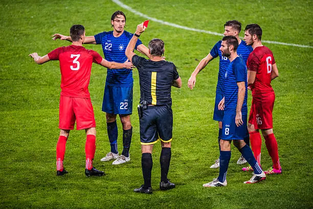 Referee holds up red card during football match, players arguing with referee.