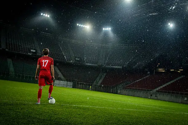 Rear view of football player standing with football on football pitch in stadium.