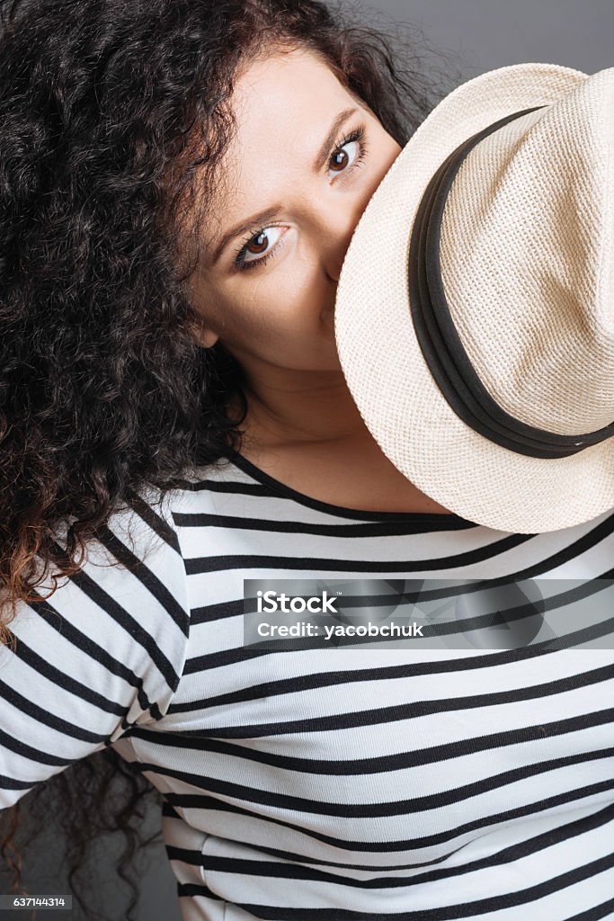 Close up of enigmatical female covering her lips I am looking at you. Positive delighted curly brunette wearing striped jumper keeping white hat near eyes while looking at camera Adult Stock Photo