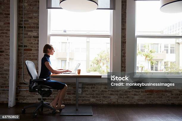 Businesswoman Sitting At Desk By Window Working On Laptop Stock Photo - Download Image Now