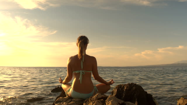 CLOSE UP: Healthy woman meditating on rocky ocean shore at magical golden sunset