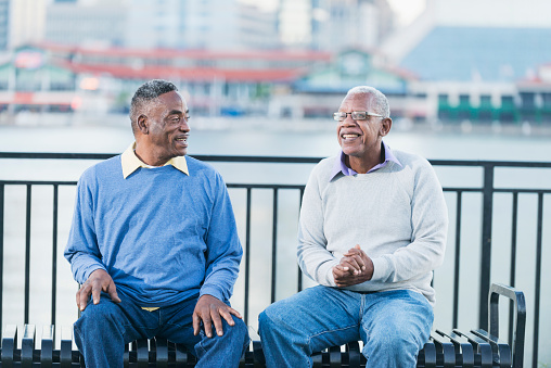 Two senior African American men sitting together on a bench by a city waterfront. They are retired, hanging out, wearing casual clothing and smiling as they chat.
