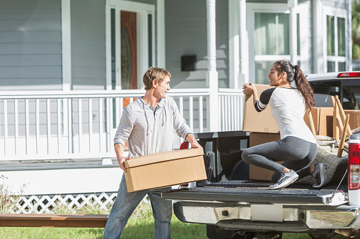 A young couple moving into a house or apartment. Their pickup truck is full of cardboard boxes and furniture. They are smiling, lifting the boxes. The woman is mixed race Hispanic and Pacific Islander ethnicity.