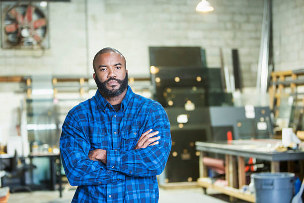 African American man in a workshop An African American man in his 30s looking at the camera with a serious expression, arms folded. He is a worker in a factory workshop, worktable and tools out of focus in the background. man beard plaid shirt stock pictures, royalty-free photos & images