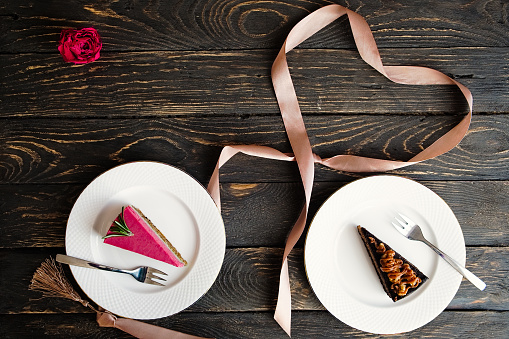 Valentine's day desserts. Heart shaped ribbon, two raspberry and chocolate cake slices on white plates. Dark wooden table