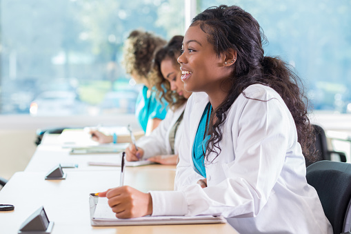 Confident African American medical student smiles while taking note in spiral notebook during class. She has long black hair and is wearing a lab coat.
