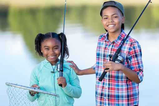 Cheerful African American brother and sister prepare to fish together. The girl is holding a fishing pole and fishing net. Her brother has his hand on her shoulder and is holding a fishing pole.