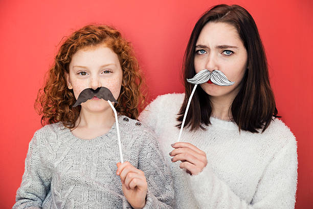 Woman and girl making faces with props mustaches, red wall. Young woman and little girl making faces with props mustaches on red wall. They are holding paper mustaches glue to a straw. Little girl has bright red hair, young woman dark brown hair. Both are wearing will sweaters. Horizontal waist up studio shot with copy space. This was shot in Quebec, Canada. women movember mustache facial hair stock pictures, royalty-free photos & images