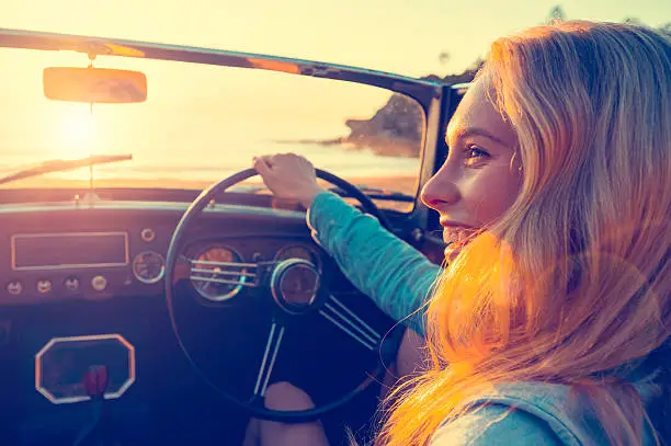 Woman driving a convertible at the beach. She is happy and smiling.  It is sunset or sunrise. With the ocean in the background. Can be flipped for left hand drive car.