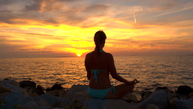 CLOSE UP: Beautiful woman in lotus yoga position by the ocean on magical evening