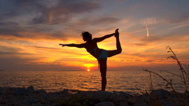CLOSE UP: Flexible young woman doing yoga king dancer pose on rocky ocean shore