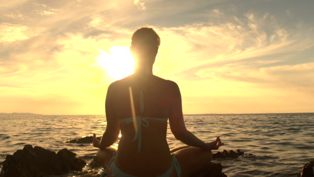 CLOSE UP: Young woman in lotus yoga position by the ocean on magical evening
