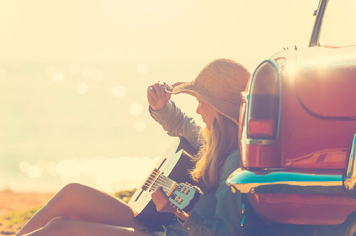 Woman with guitar leaning on a car. The car is parked at the beach at sunrise. Very relaxing vacation or road trip image. Car is a convertible. Focus on background. copy space