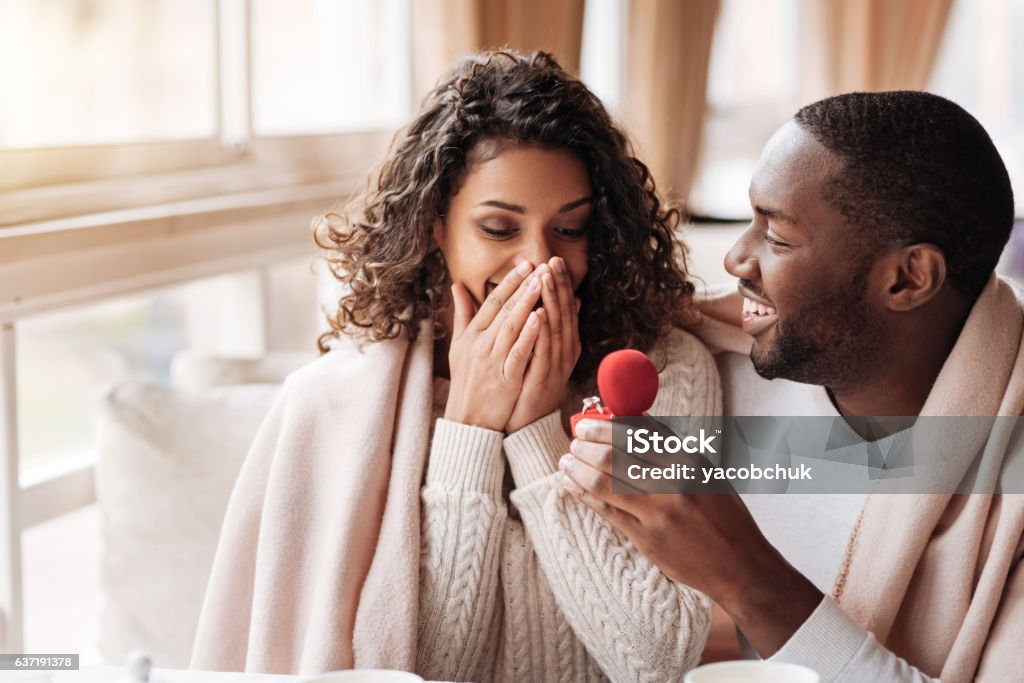 Amazed African American couple getting engaged in the cafe I love you. Amazed surprised positive African American couple sitting in the cafe and being covered with a blanket while getting engaged Engagement Stock Photo