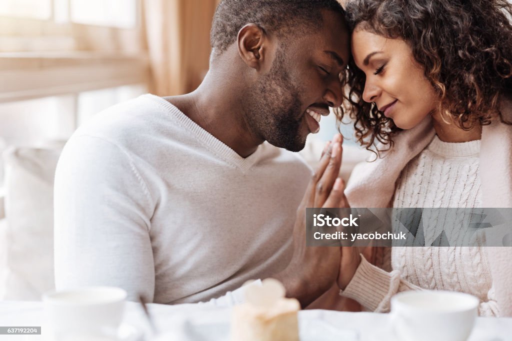 Passionate African American couple touching hands in the cafe Peacefulness in us. Delighted peaceful positive African American couple sitting in the cafe and touching hands of each other while expressing peacefulness and love Couple - Relationship Stock Photo