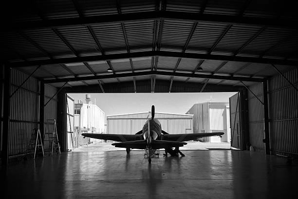 Silhouette of a P-51 Mustang fighter aircraft in a hangar A P-51 Mustang aircraft is prepared for flight in a hangar at Archerfield aerodrome in Brisbane, Queensland, Australia. The photo shows the silhouette of the fighter warbird aeroplane against the bright backdrop of other hangars outside. p51 mustang stock pictures, royalty-free photos & images