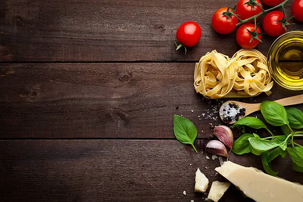 Ingredients for cooking traditional italian pasta shot on rustic wood table. The ingredient are paced at the right side of the frame leaving a useful copy space at the center-left side. Composition includes tagliatelle pasta, olive oil, tomatoes, basil, pepper, salt, garlic and parmesan cheese. Low key DSRL studio photo taken with Canon EOS 5D Mk II and Canon EF 100mm f/2.8L Macro IS USM