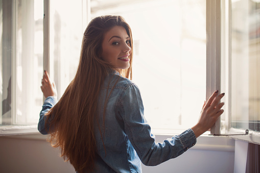 Rear view of a beautiful young woman opening the window, looking over the shoulder and smiling