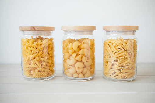 three transparent pots with wooden tops full of different kinds of raw pasta on light grey background. 