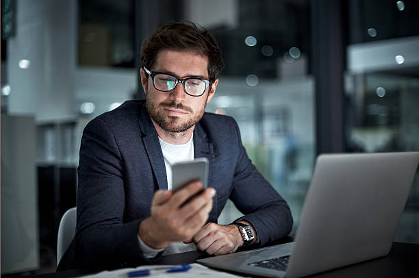 Enhancing his entrepreneurial ambition with the right tools Shot of a young businessman using his laptop and phone at work guy looking at cell phone stock pictures, royalty-free photos & images