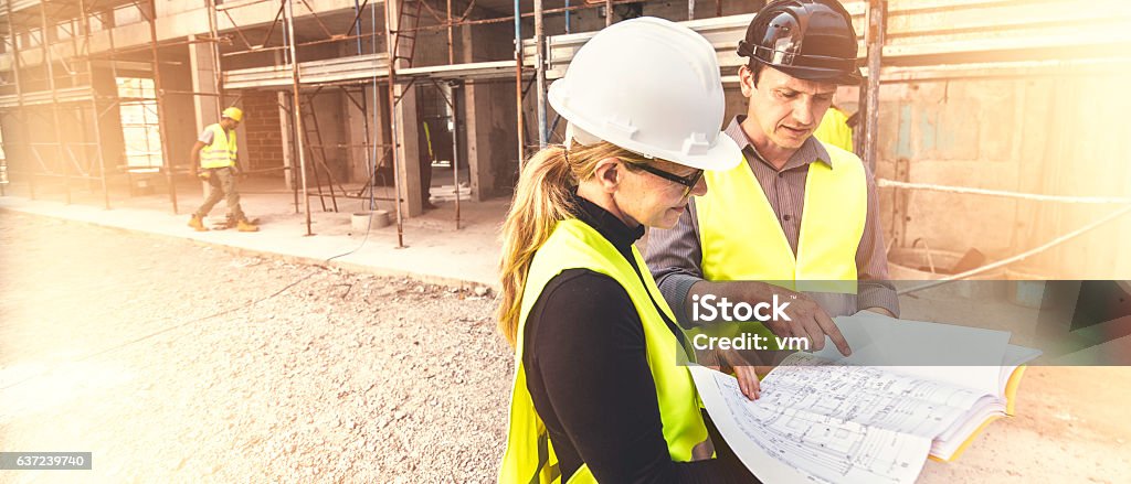 Reviewing blueprints on a construction site A man and a woman in a reflective clothing standing at a construction site and examining blueprints, scaffolding in background. Construction Industry Stock Photo