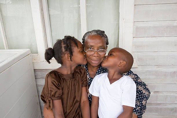 Series:Proud Honduran grandmother getting kiss from grandchildren Portrait of senior Honduran woman in her sixties with her young granddaughter and grandson. They live on the island of Roatan in a shanty town. She is sitting with her arms around them and they are giving her a kiss on the cheek. They are outside their house. There is a washing machine on the front porch. Taken with Canon 5D Mark lll. poverty stock pictures, royalty-free photos & images