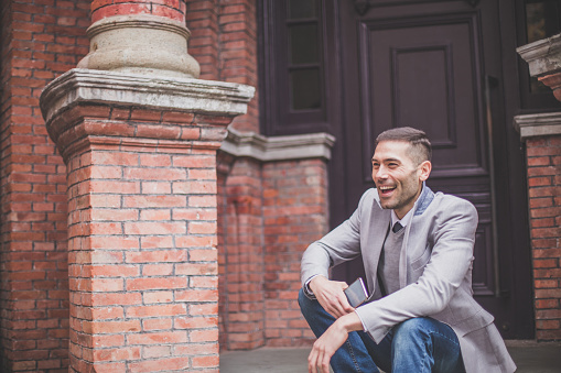 Businessman dressed in suit sitting on the doorstep with a mobile phone in his hand, waiting for a friend to come.