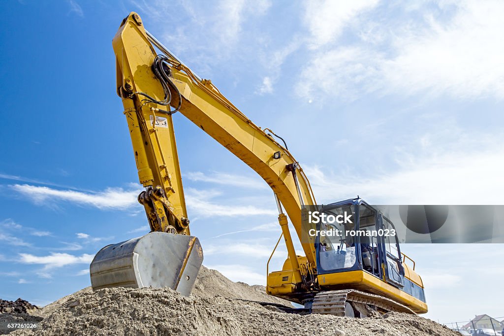 Excavator is preparing pile of sand for loading in truck. Yellow excavator is making pile of soil by pulling ground up on heap at construction site, project in progress. Backhoe Stock Photo