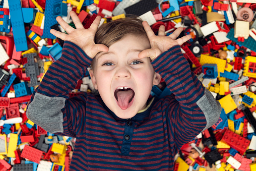 A little happy blond boy with blond hair and blue eyes lies between a lot of colorful plastic blocks toy / building blocks.