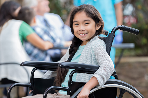 Cheerful Filipino girl smiles after receiving care at an outdoor medical clinic. She is sitting in a wheelchair and smiling at the camera. Doctors, nurses and patients are in the background.
