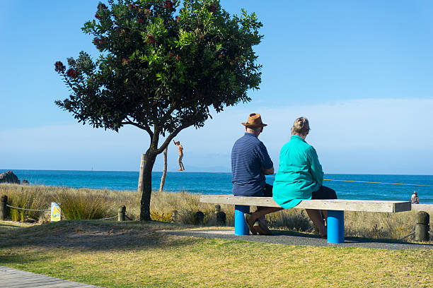 Mount Maunganui, New Zealand - December 21, 2016: Elderly couple rest and look at scenic ocean view on bench seat along Mount Maunganui ocean-beach while man appears to be walking above the water beyond beach while practicing the sport of slack-lining