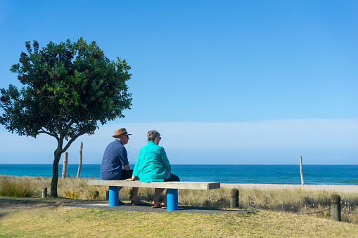 Mount Maunganui, New Zealand - December 21, 2016: Elderly couple rest sitting together looking at scenic ocean view on bench seat along Mount Maunganui ocean-beach