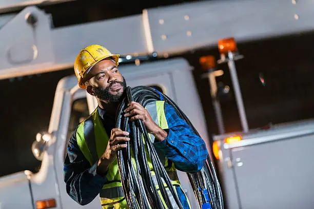 An African American man wearing a hard hat and safety vest, standing in front of a bucket truck or cherry picker, carrying cables on his shoulder. He is a construction worker, utility worker or engineer.