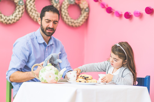 An adorable little girl is having a tea party with her father and her imaginary friend stuffed animals  in a bright pink room