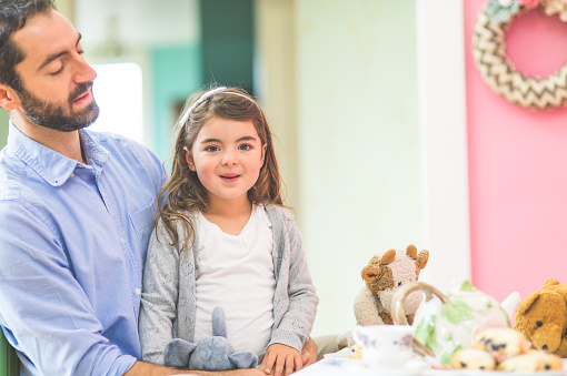 An adorable little girl is having a tea party with her father and her imaginary friend stuffed animals  in a bright pink room. She is looking at the camera and smiling