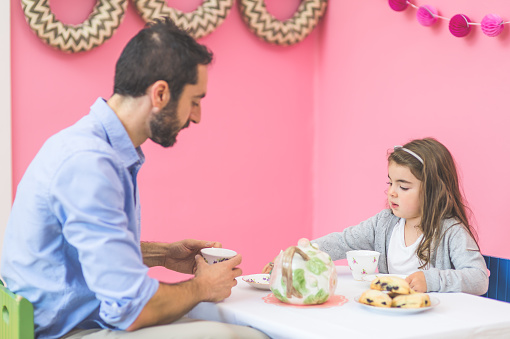 An adorable little girl is having a tea party with her father and her imaginary friend stuffed animals  in a bright pink room