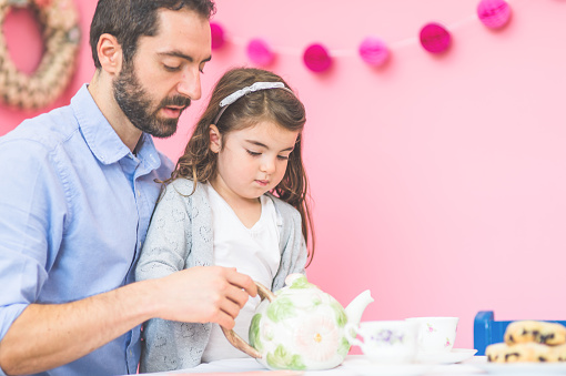 An adorable little girl is having a tea party with her father and her imaginary friend stuffed animals  in a bright pink room. They are pouring tea together