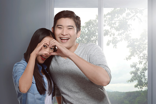 Happy asian couple in love gesturing a heart with fingers with windows background