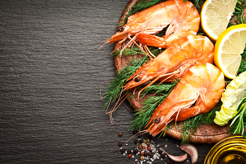 Top view of three king prawns on a wooden plate with some cooking and seasoning ingredients like salt, pepper, olive oil, garlic, lime, and rosemary at the right side of a dark slate background leaving a useful copy space at the center-left of the frame. DSRL studio photo taken with Canon EOS 5D Mk II and Canon EF 100mm f/2.8L Macro IS USM