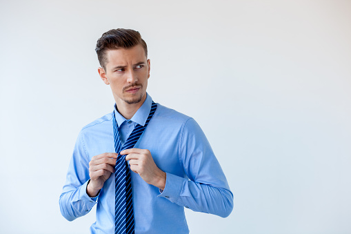 Portrait of suspicious young Caucasian businessman wearing blue shirt knotting his tie and looking away